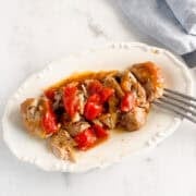 Cooked chicken pieces with tomatoes and sauce on a white plate, next to a fork and a blue napkin.