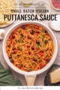 A pan of spaghetti puttanesca with tomato sauce, olives, and herbs sits on a countertop next to uncooked pasta, grated cheese, and seasonings. Text overlay reads Small Batch Italian Puttanesca Sauce.