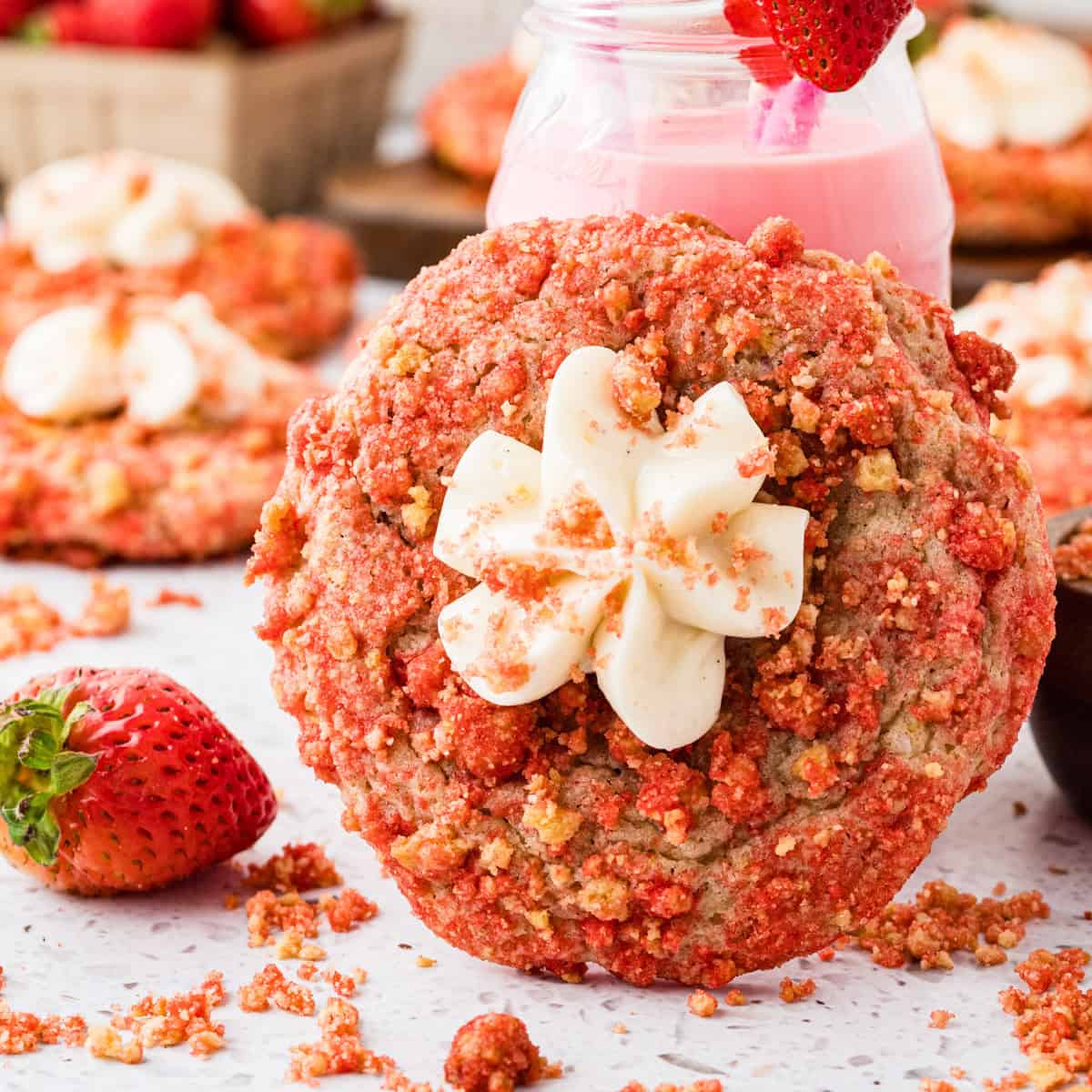 A strawberry crumble cookie topped with a swirl of cream cheese frosting, surrounded by fresh strawberries and cookie crumbs, with a jar of pink milk in the background.