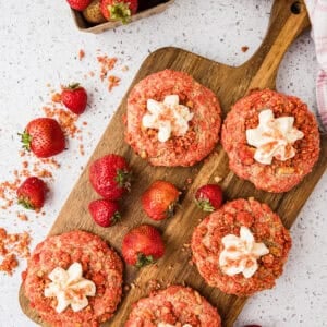 Six strawberry crunch cookies, some topped with white frosting, are arranged on a wooden board with fresh strawberries. A container of strawberries and a pink towel are nearby on a white countertop.