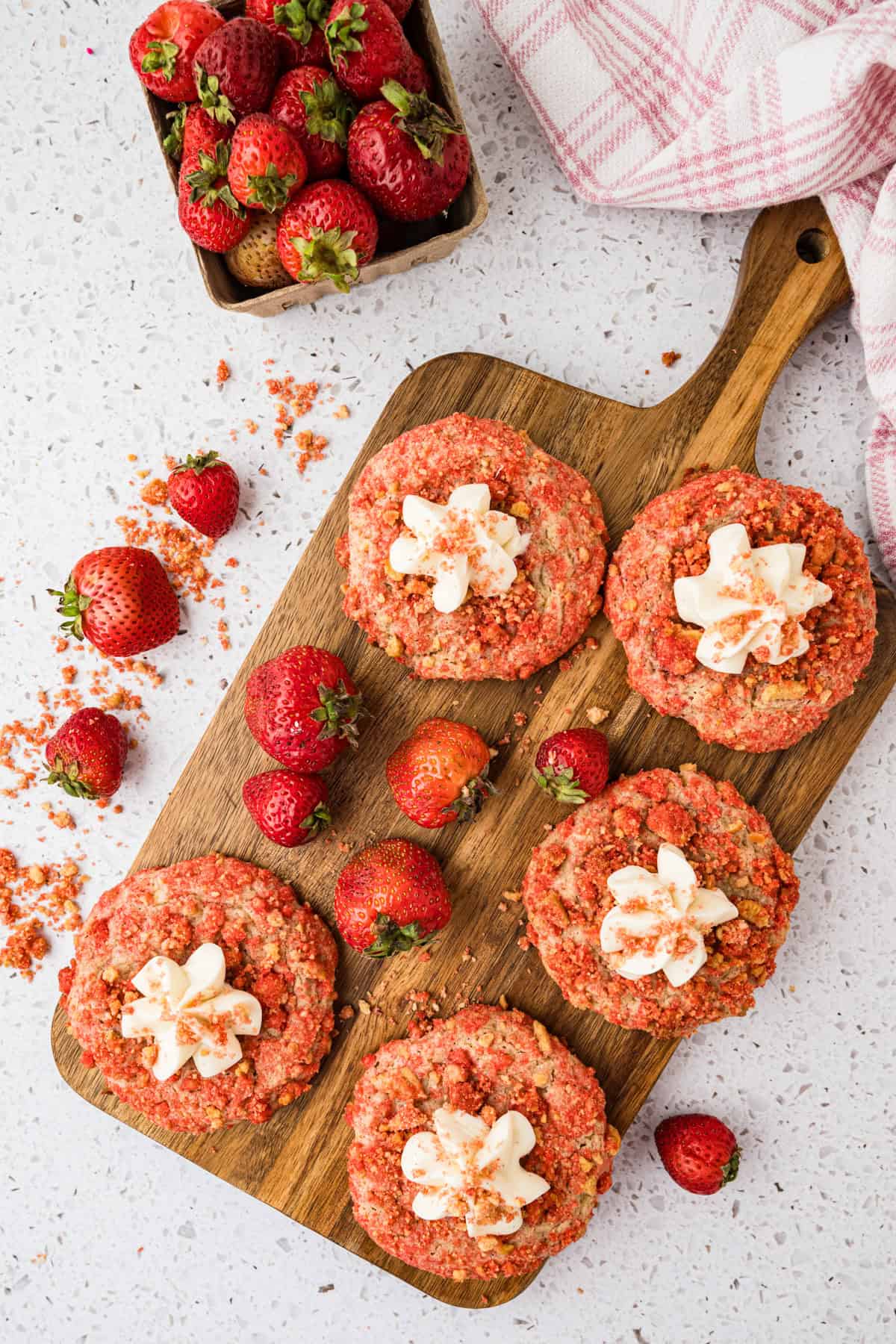 Six strawberry crunch cookies, some topped with white frosting, are arranged on a wooden board with fresh strawberries. A container of strawberries and a pink towel are nearby on a white countertop.