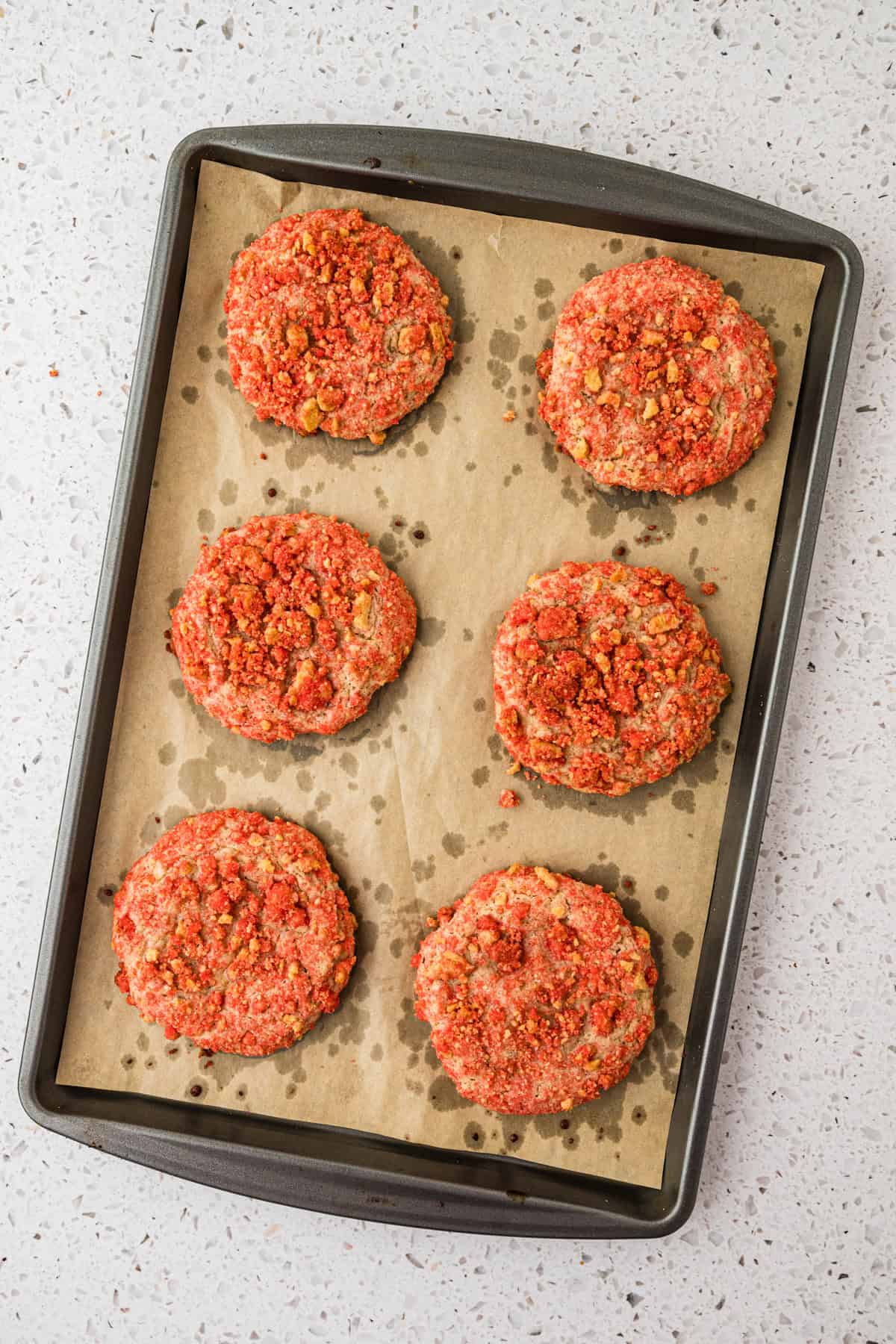 Six round, crumb-coated lemon crinkle cookies with a reddish topping are arranged on a parchment-lined baking sheet atop a light speckled surface.