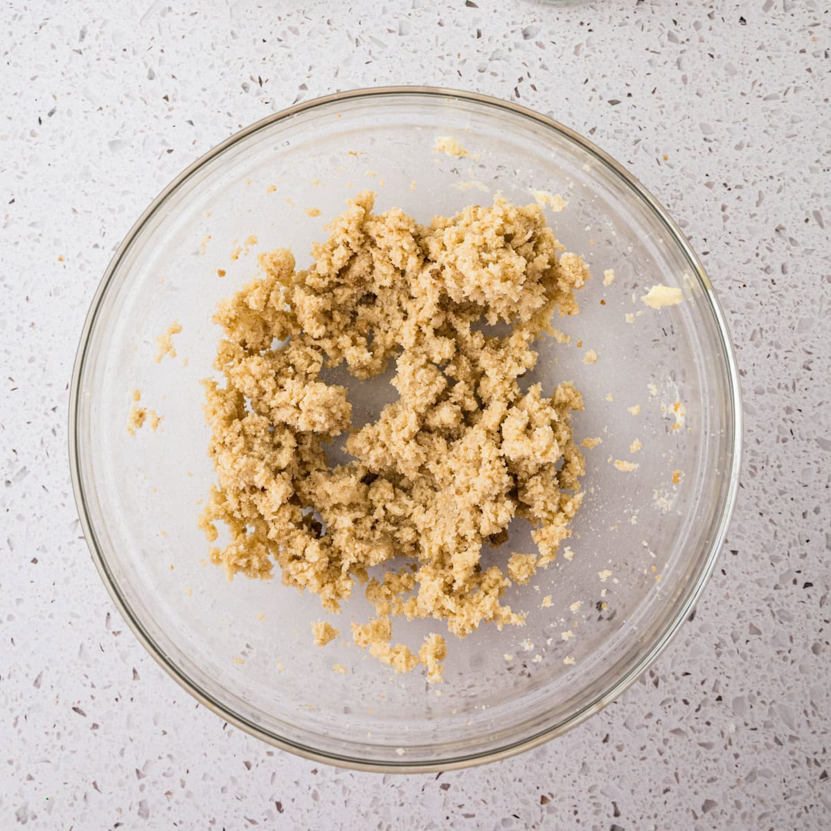 A glass bowl containing a coarse, light brown mixture for strawberry crunch cookies sits on a speckled white countertop.