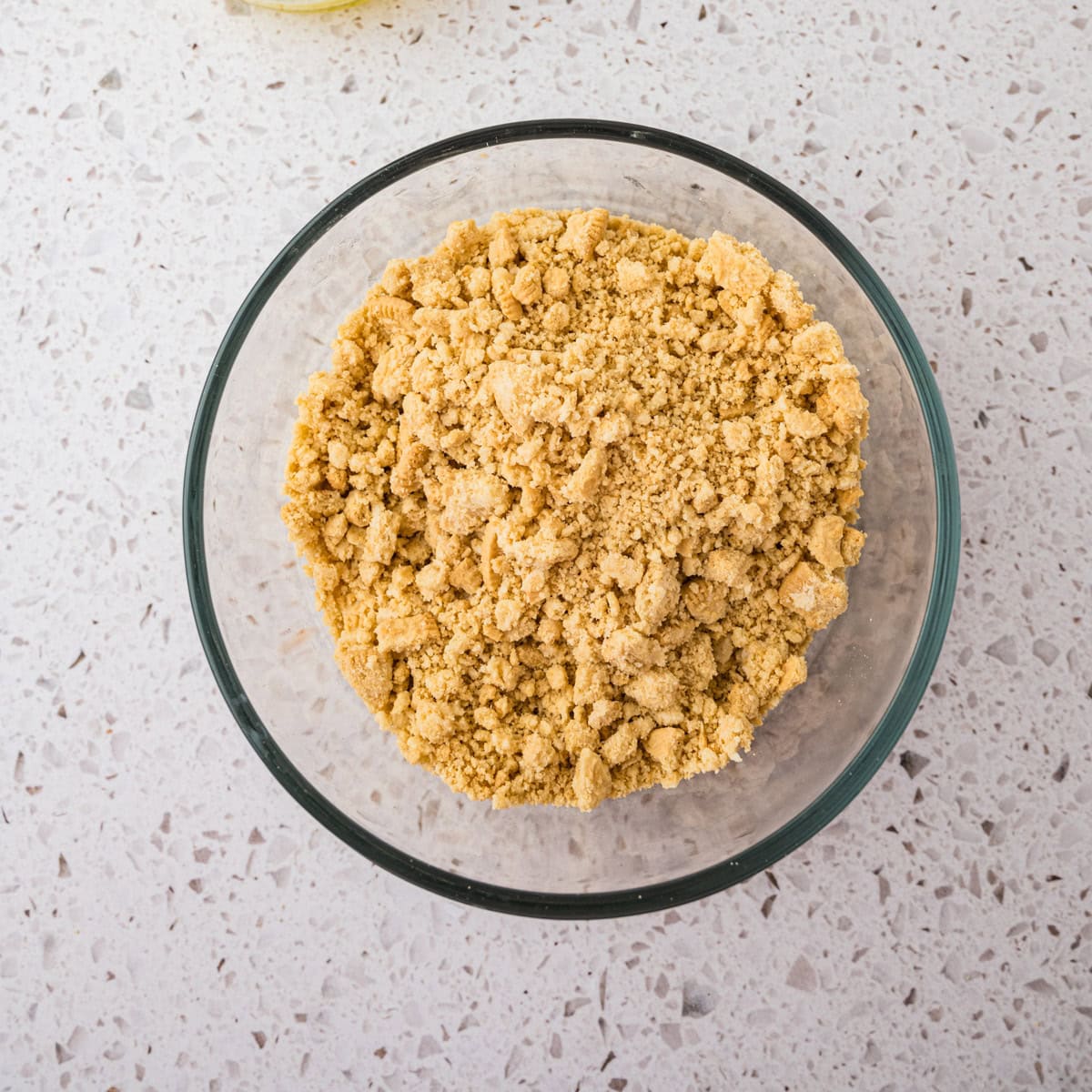 A glass bowl filled with crushed golden oreo crumbs sits on a light-colored speckled countertop, ready to be used in a recipe for lemon crinkle cookies.