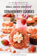 A large strawberry crunch cookie with cream topping sits in front of a pink milkshake, fresh strawberries, and more cookies on a white surface. Text reads, “Small Batch Crunchy Strawberry Cookies.”.