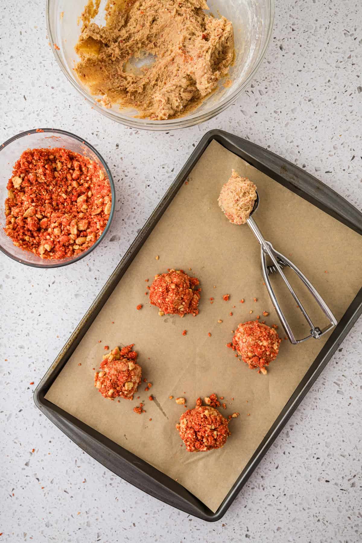 A baking tray with four unbaked lemon crinkle cookies coated in red crumbs, a bowl of red crumb mixture, a bowl of raw dough, and a cookie scoop on a speckled countertop.