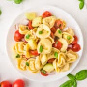 A plate of tortellini pasta salad with cherry tomatoes, cucumber, red onion, and fresh basil, viewed from above on a white surface.
