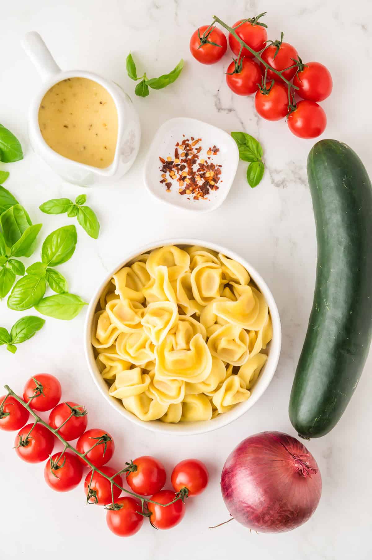 A bowl of tortellini surrounded by fresh basil, cherry tomatoes on the vine, a cucumber, a red onion, vinaigrette, and a dish of red pepper flakes on a white surface.