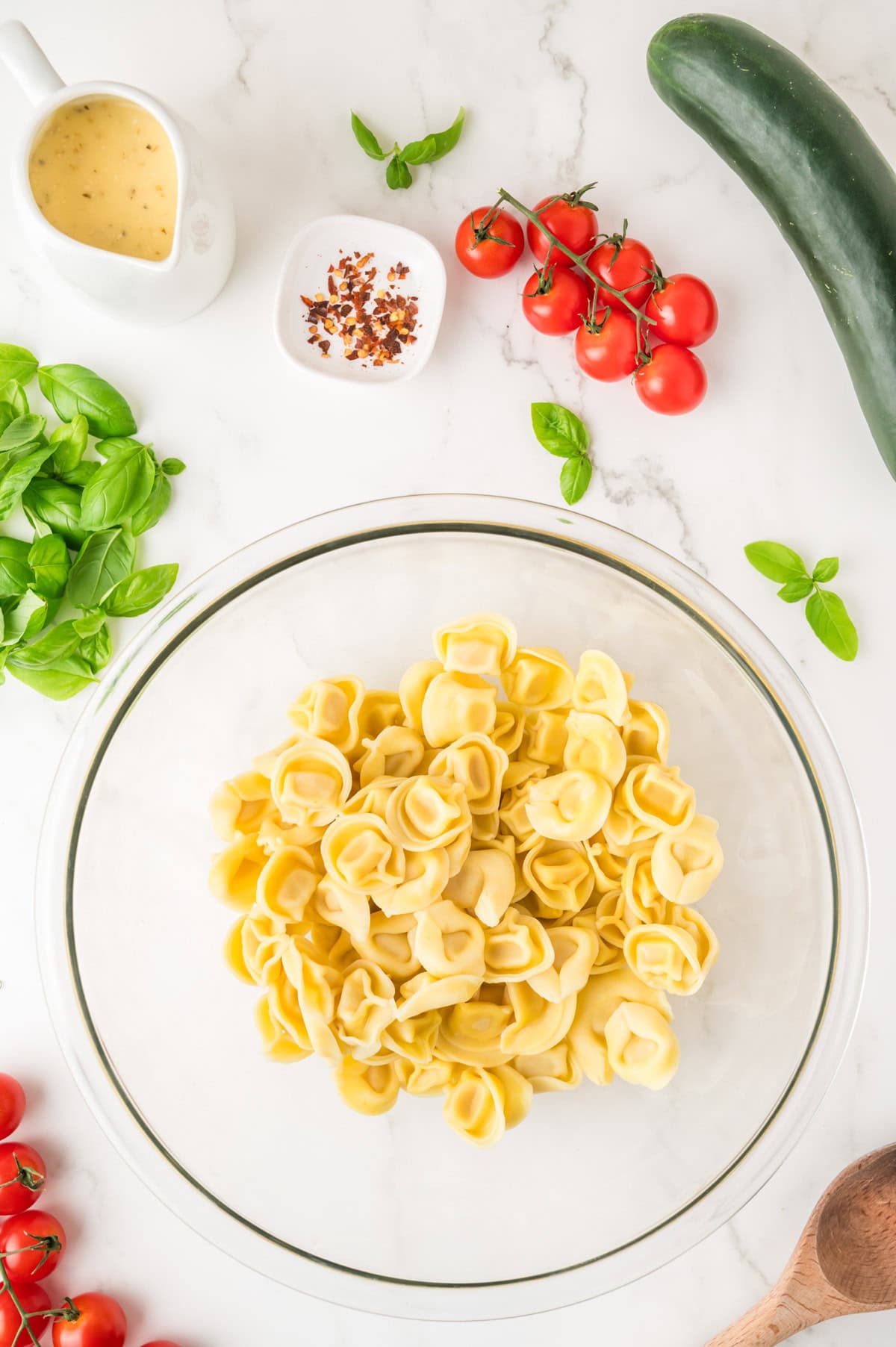 A glass bowl of tortellini surrounded by fresh basil, cherry tomatoes on the vine, a cucumber, red pepper flakes, and a small pitcher of dressing on a white surface.