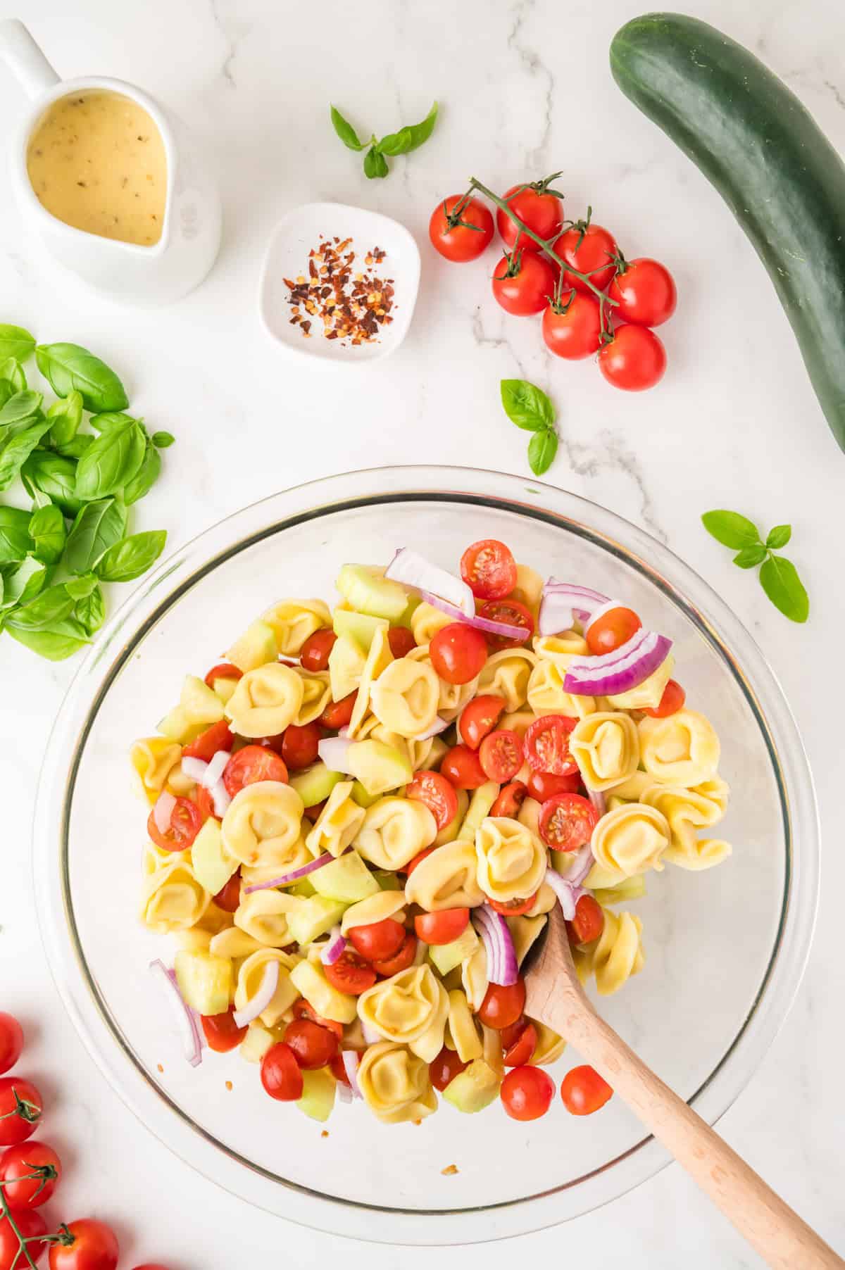 A glass bowl of tortellini pasta salad with cherry tomatoes, red onion, cucumber, and basil on a white surface, surrounded by fresh ingredients and a small jug of dressing.