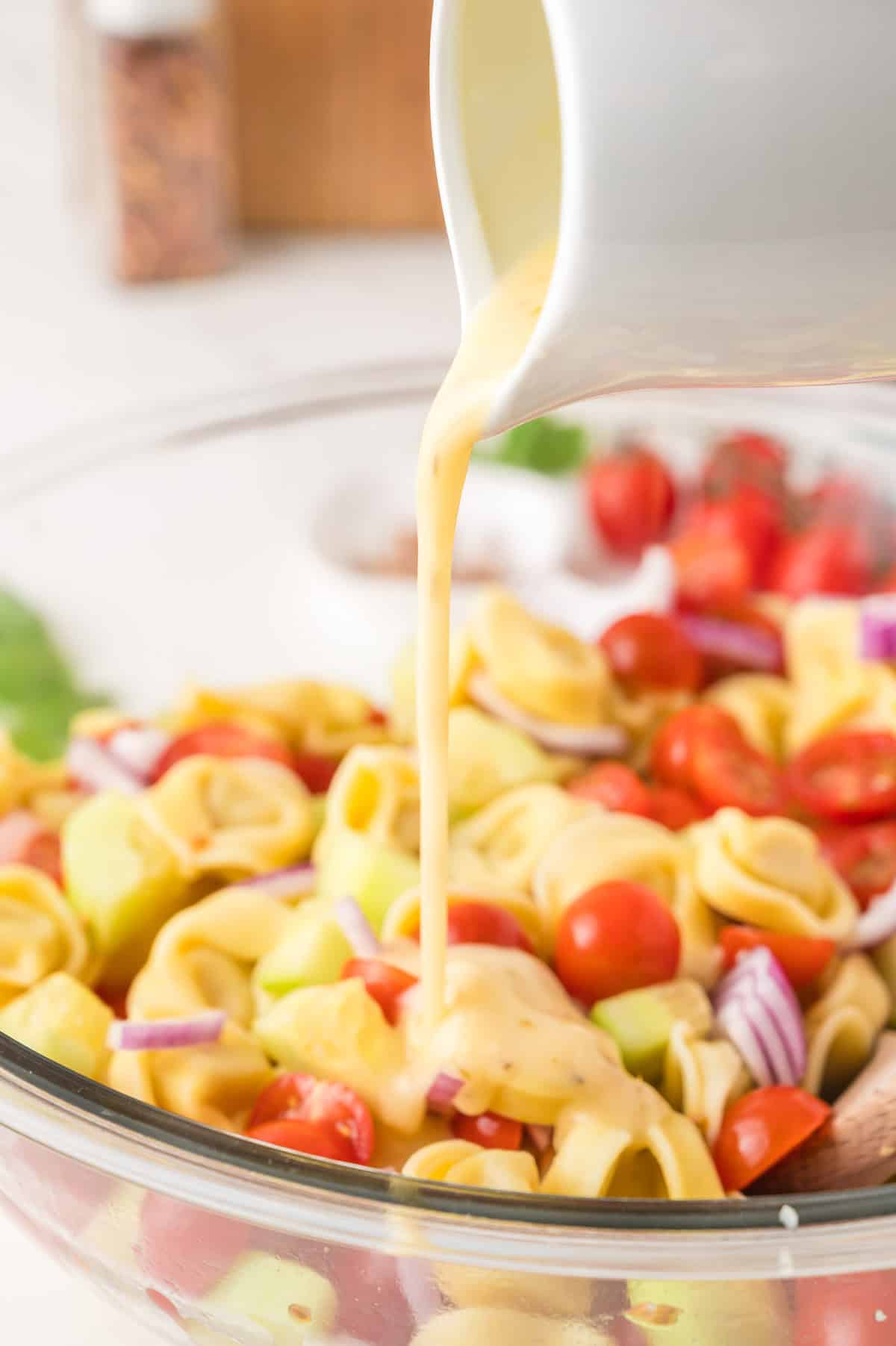 Cheese tortellini, cherry tomatoes, red onion, and cucumber in a glass bowl with creamy dressing being poured on top.