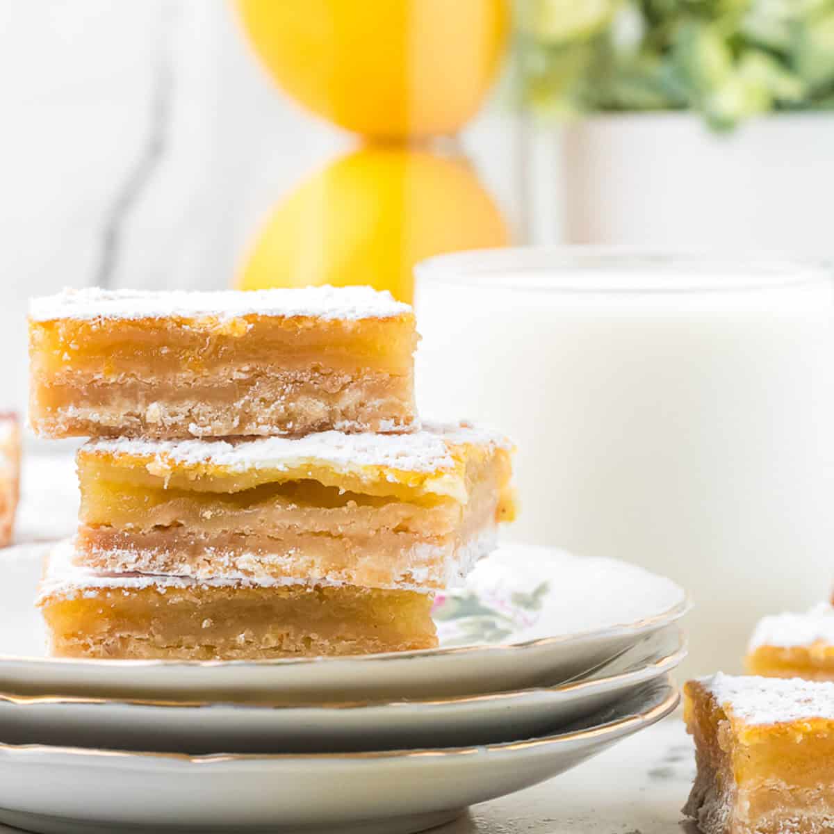 Three lemon bars dusted with powdered sugar are stacked on plates in front of a glass of milk, with blurred decor in the background.