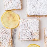 Four lemon bars dusted with powdered sugar are arranged on a white surface with two lemon slices nearby.