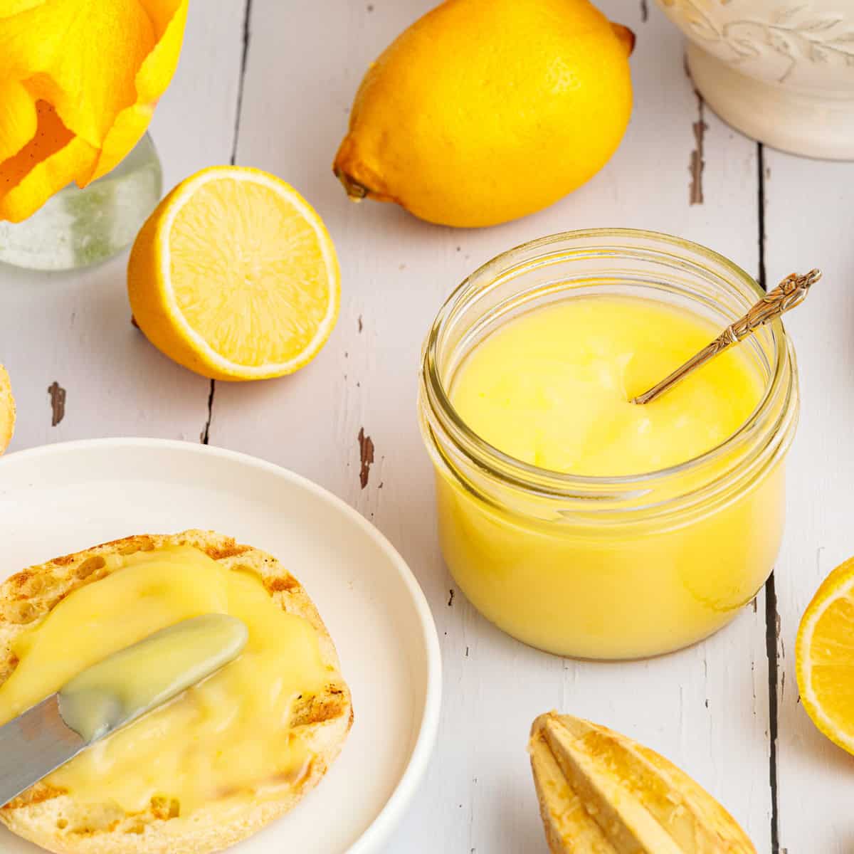 A jar of lemon curd with a spoon, surrounded by lemons, a halved lemon, and a buttered English muffin on a white plate.