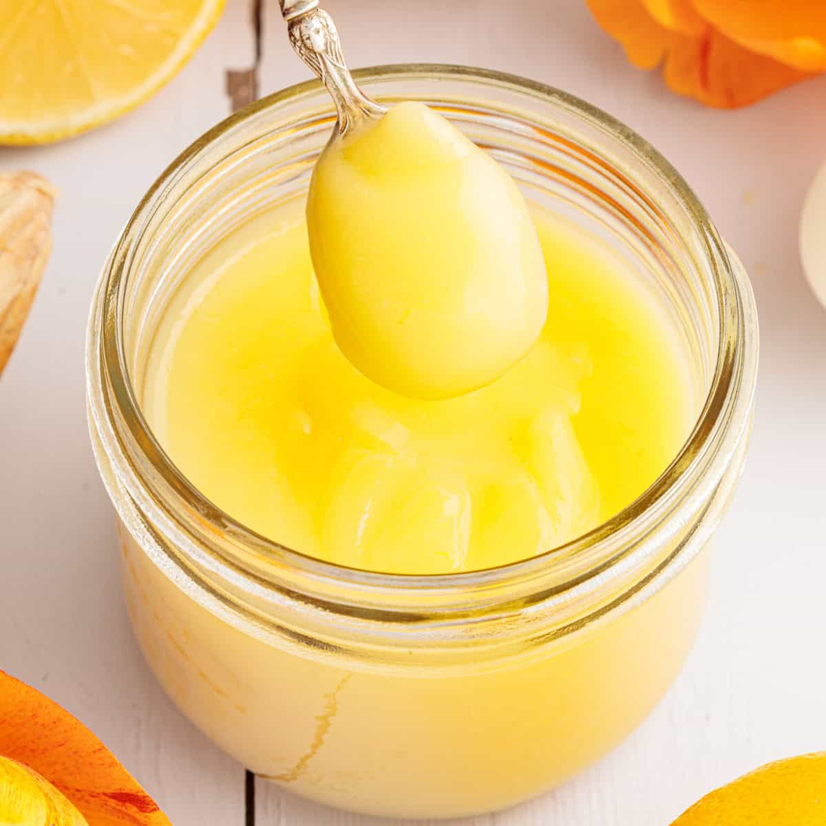A jar of lemon curd with a spoonful being lifted out, surrounded by pieces of lemon and orange on a white surface.
