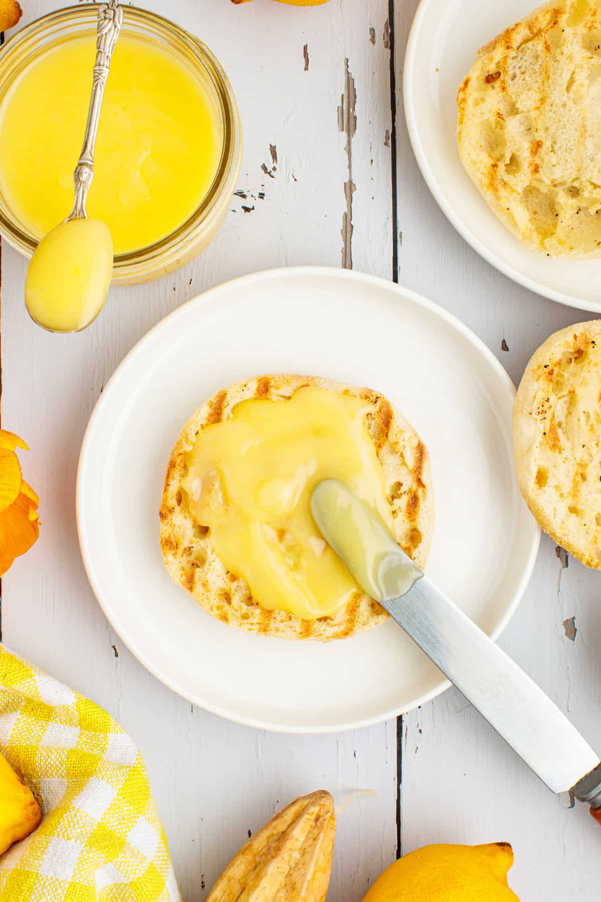 An English muffin half spread with lemon curd sits on a white plate, next to a knife, a jar of lemon curd, and more muffins on a light wooden table.