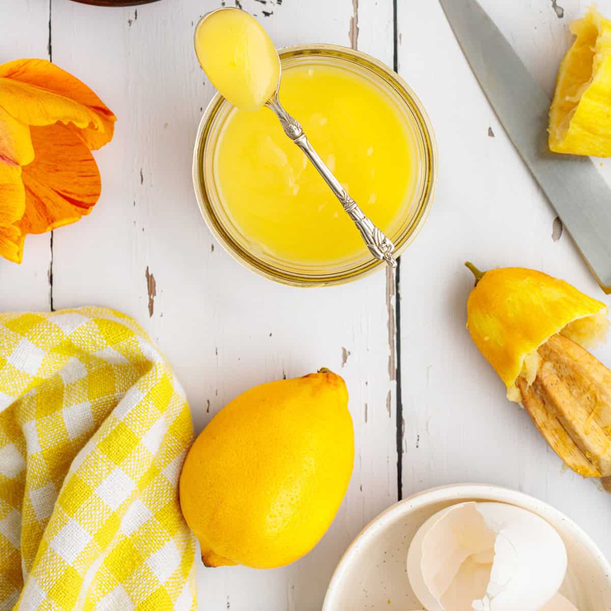 A jar of lemon curd with a spoon, a whole lemon, lemon peel, eggshells, a yellow checkered cloth, and a knife on a white wooden surface.