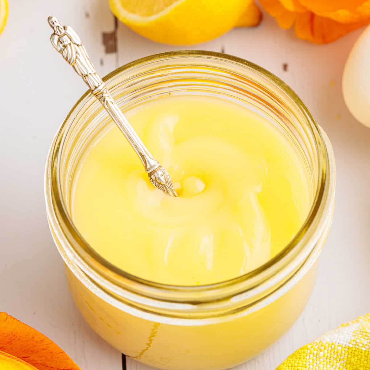 A glass jar filled with smooth yellow lemon curd, with a decorative silver spoon inserted, surrounded by lemon slices and a white surface.