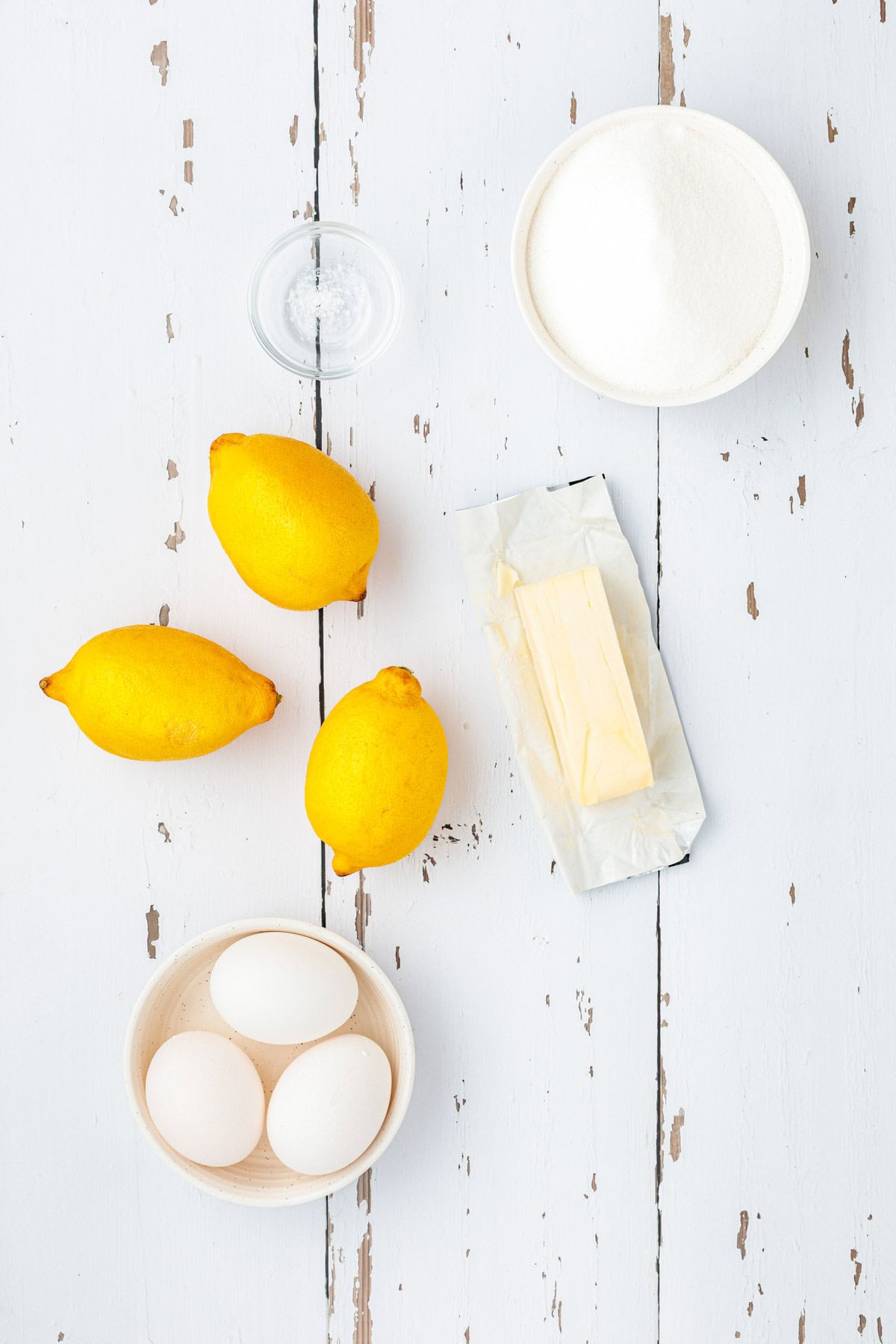 Three lemons, three eggs, a glass of water, a bowl of sugar, and two sticks of butter on a white wooden surface.