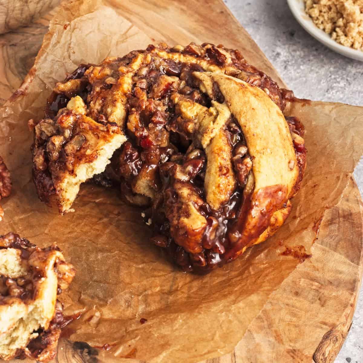 A sticky pecan cinnamon roll with a bite taken out, sitting on brown parchment paper on a wooden board. Crumbles are visible in the background.
