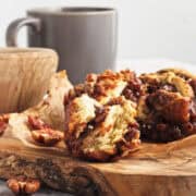 Cinnamon roll with chopped pecans and caramel on a wooden board, next to a mug and wooden bowl.