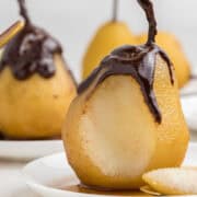 A poached pear with chocolate sauce drizzled on top sits on a white plate, with more pears and sauce in the background.