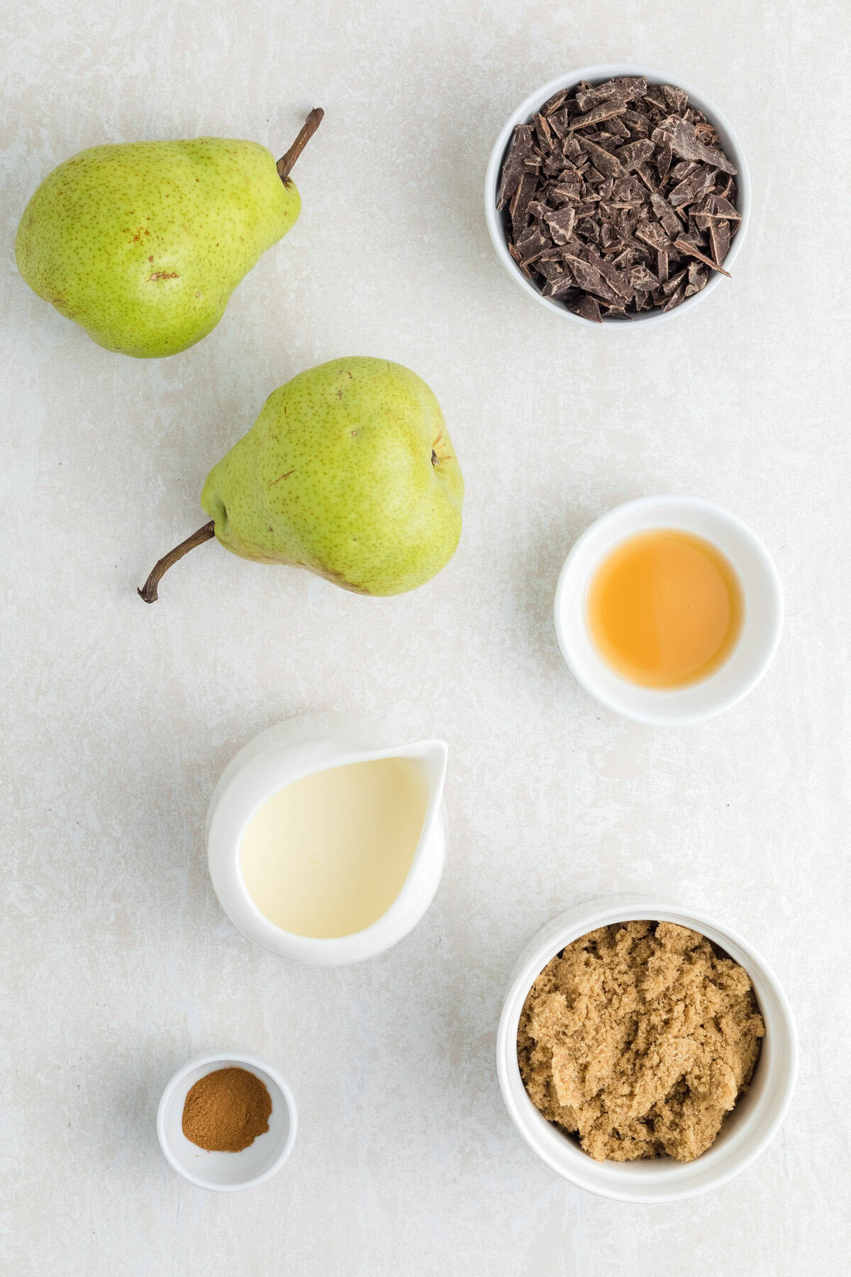 Two green pears, a bowl of chopped chocolate for chocolate sauce, a bowl of brown sugar, a small jug of cream, vanilla extract, and a pinch of ground cinnamon rest on a light surface.