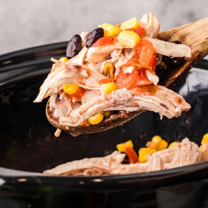 A wooden spoon holds shredded chicken, black beans, corn, and diced tomatoes over a slow cooker containing the same mixture.