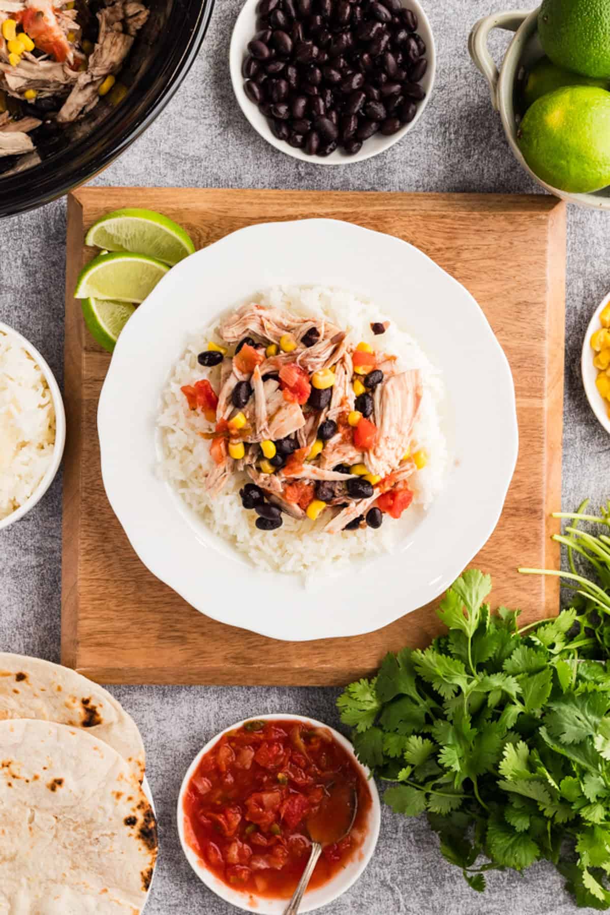 A plate of white rice topped with shredded meat, black beans, corn, and salsa is surrounded by tortillas, salsa, cilantro, lime, and other ingredients on a wooden board.