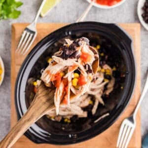 A wooden spoon holds a serving of shredded chicken, black beans, corn, and salsa over a slow cooker, surrounded by bowls of rice, salsa, and greens.