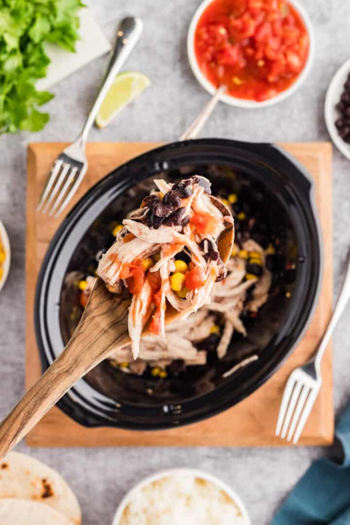 A wooden spoon holds a serving of shredded chicken, black beans, corn, and salsa over a slow cooker, surrounded by bowls of rice, salsa, and greens.
