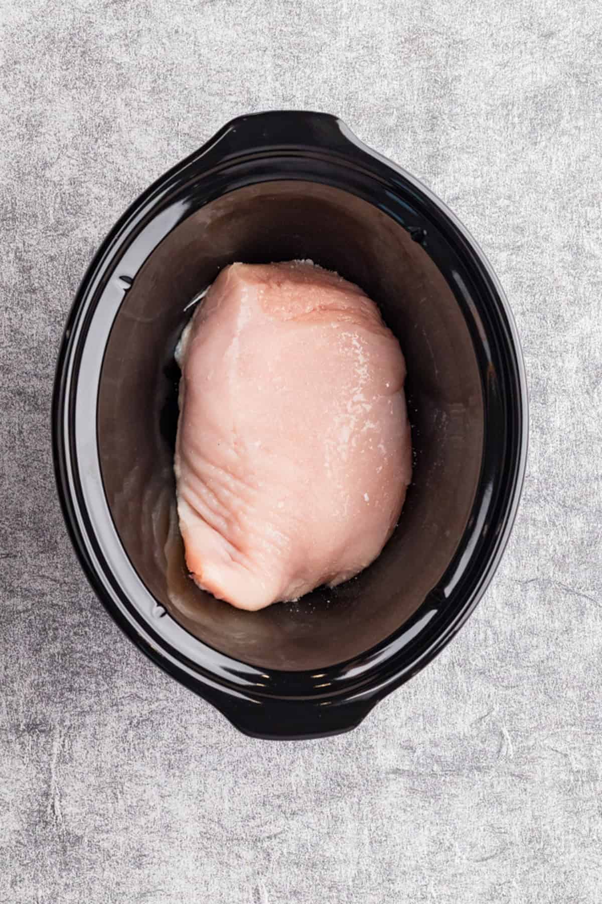 Raw chicken breast placed in a black slow cooker on a gray textured surface.