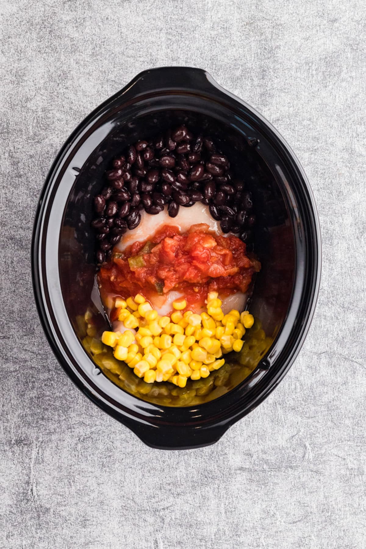 A slow cooker filled with raw chicken breast, black beans, corn, and salsa arranged in separate sections on top.