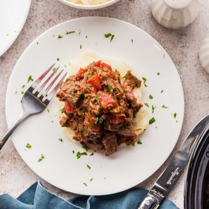 A plate of braised beef with tomato sauce served over mashed potatoes, garnished with chopped parsley, with a fork on the side.