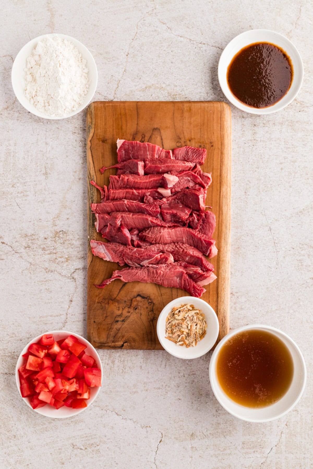 Raw beef strips on a wooden board surrounded by bowls of flour, diced tomatoes, dry seasoning, broth, and a dark sauce on a light surface.