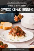 A plate of Swiss steak with vegetables served over mashed potatoes, with a fork holding a bite, bread slices, and a slow cooker in the background.