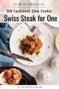 A plate of Swiss steak with tomato and herbs on mashed potatoes, next to a fork, knife, napkin, salt and pepper shakers, and a dish of mashed potatoes.