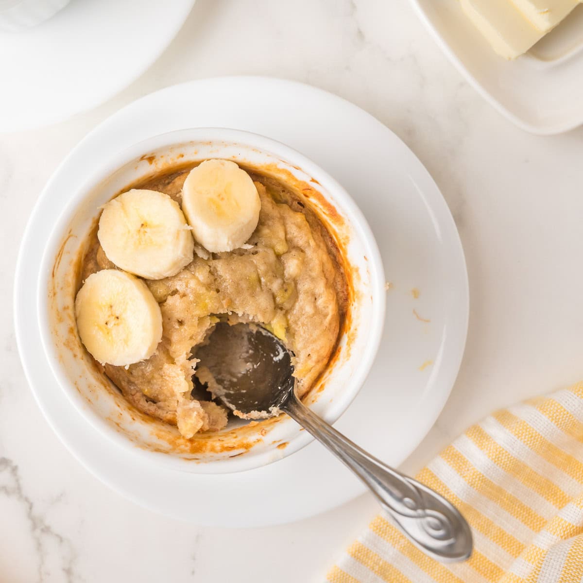 A ramekin of single serving banana bread topped with banana slices, with a spoon resting inside, on a white plate next to a striped yellow napkin.