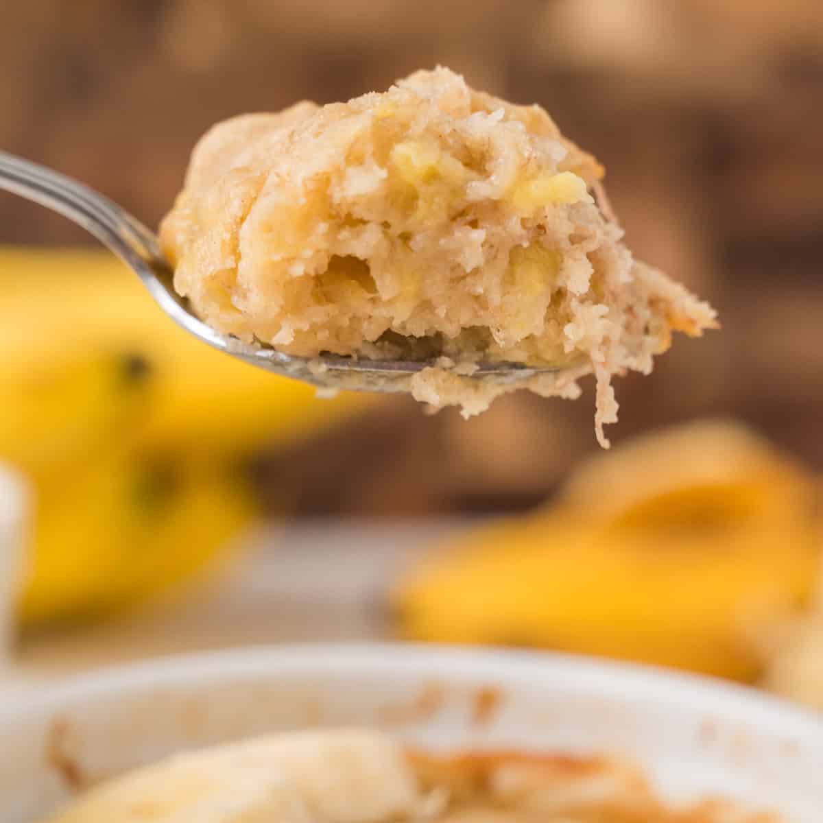 A close-up of a spoon holding a bite of banana bread with visible banana pieces, with blurred bananas in the background.
