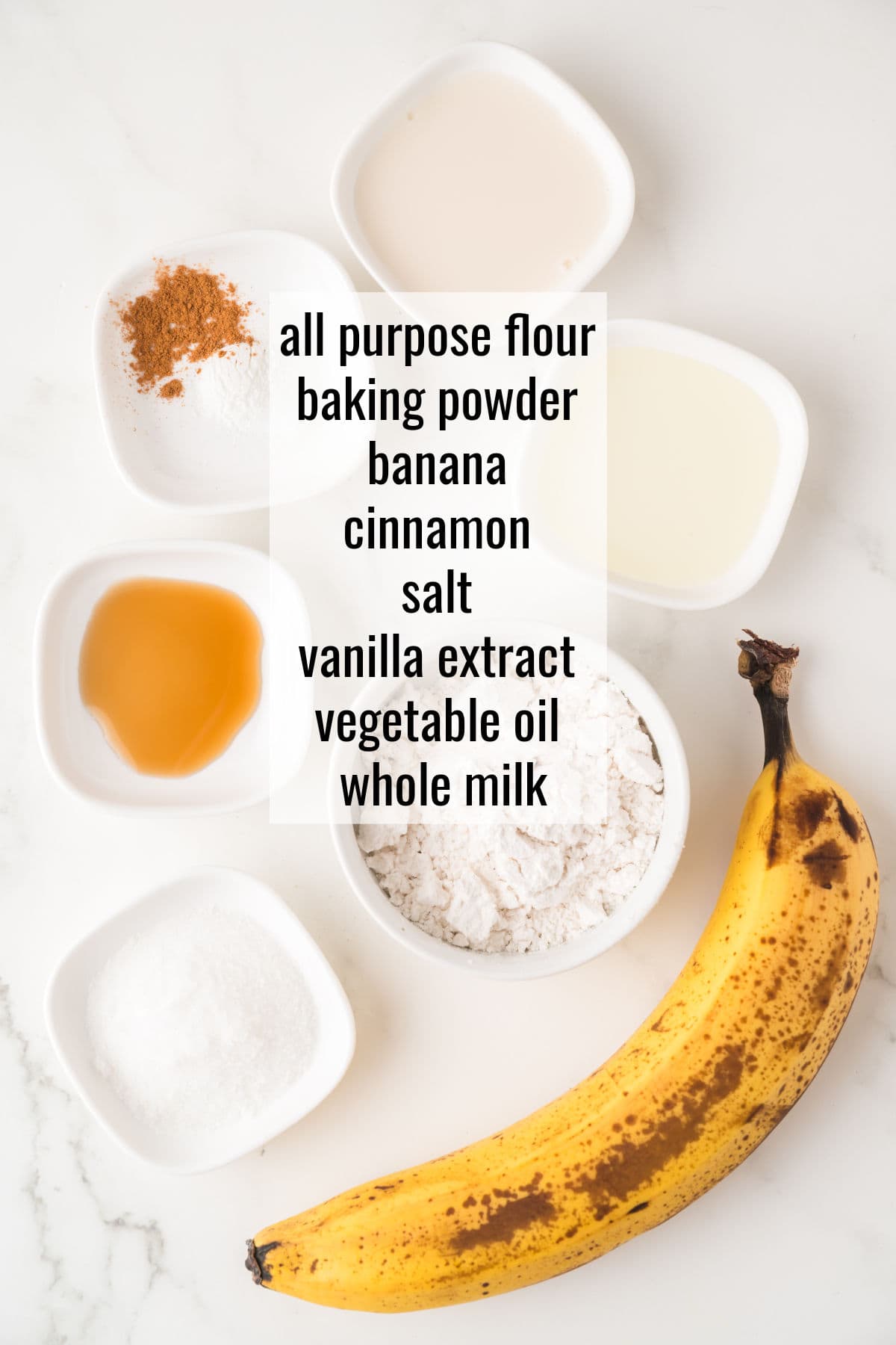 Top-down view of measured ingredients for baking, including a banana, flour, salt, cinnamon, baking powder, vanilla extract, vegetable oil, and milk, arranged on a white surface.