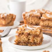 A close-up of a square slice of cinnamon coffee cake with crumb topping and white icing on a white plate, with more cake and a coffee mug in the background.