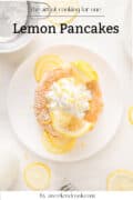 A stack of lemon pancakes topped with whipped cream and lemon slices on a white plate, surrounded by lemon slices. Text reads: Lemon Pancakes, the art of cooking for one, by aweekendcook.com.