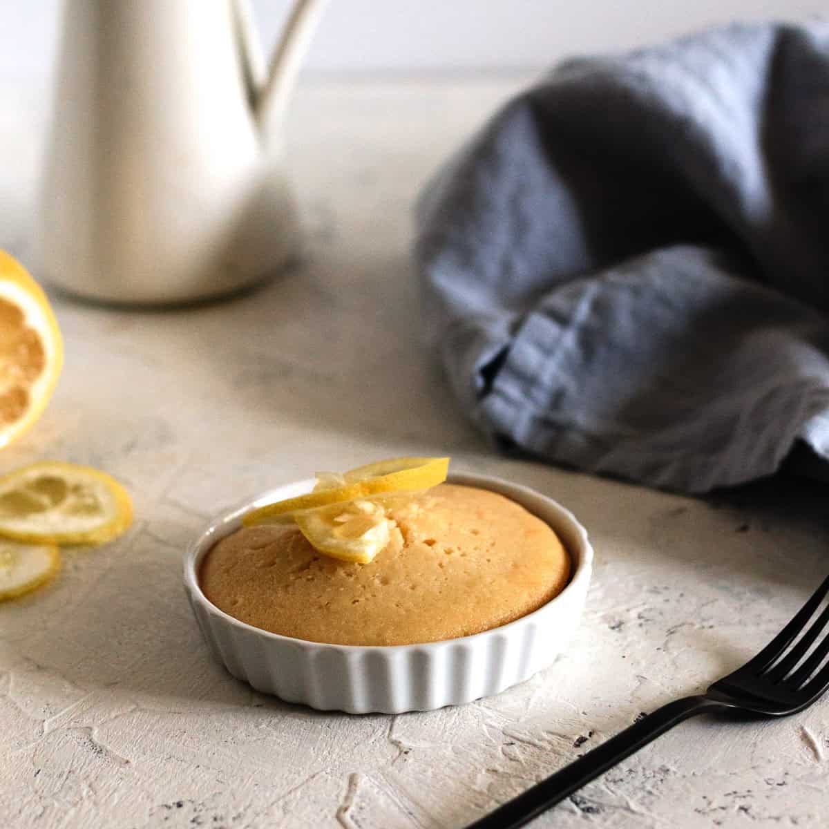 A small baked lemon cake in a white ramekin, garnished with lemon slices, sits on a textured surface near a fork, a blue napkin, a lemon, and a white pitcher.