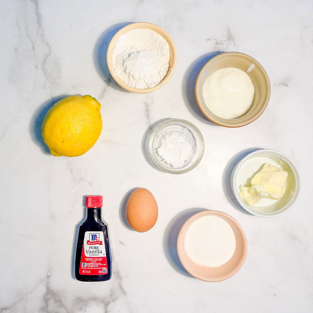 Ingredients on a marble surface: a lemon, flour, yogurt, cornstarch, butter, sugar, an egg, and a bottle of vanilla extract.