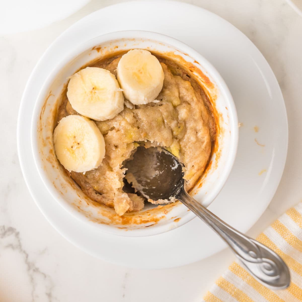 A ramekin of baked oatmeal inspired by single serving banana bread, topped with banana slices, with a spoon resting inside and a portion scooped out, sits on a white plate.