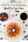 A single waffle topped with berries and whipped cream on a plate, with a cup of coffee and a bowl of mixed berries on a marble surface.