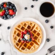 A waffle topped with whipped cream, strawberries, and blueberries on a plate, next to a bowl of mixed berries and a cup of black coffee on a marble surface.