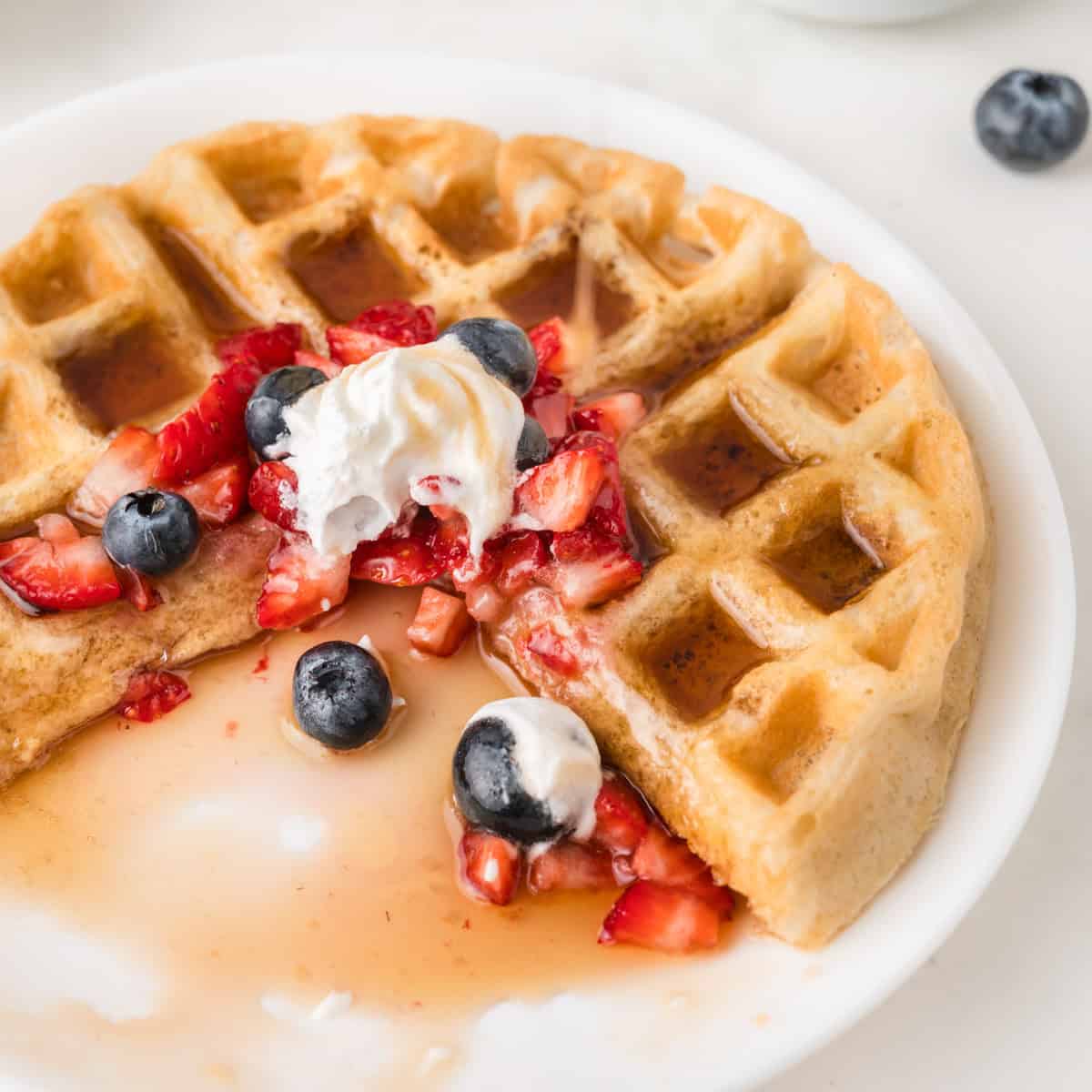 A waffle with a slice missing, topped with strawberries, blueberries, whipped cream, and syrup on a white plate.