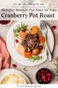 A plate of cranberry pot roast with carrots, potatoes, mashed potatoes, and garnished with rosemary and cranberries, set on a white table with a fork and a bowl of cranberries.
