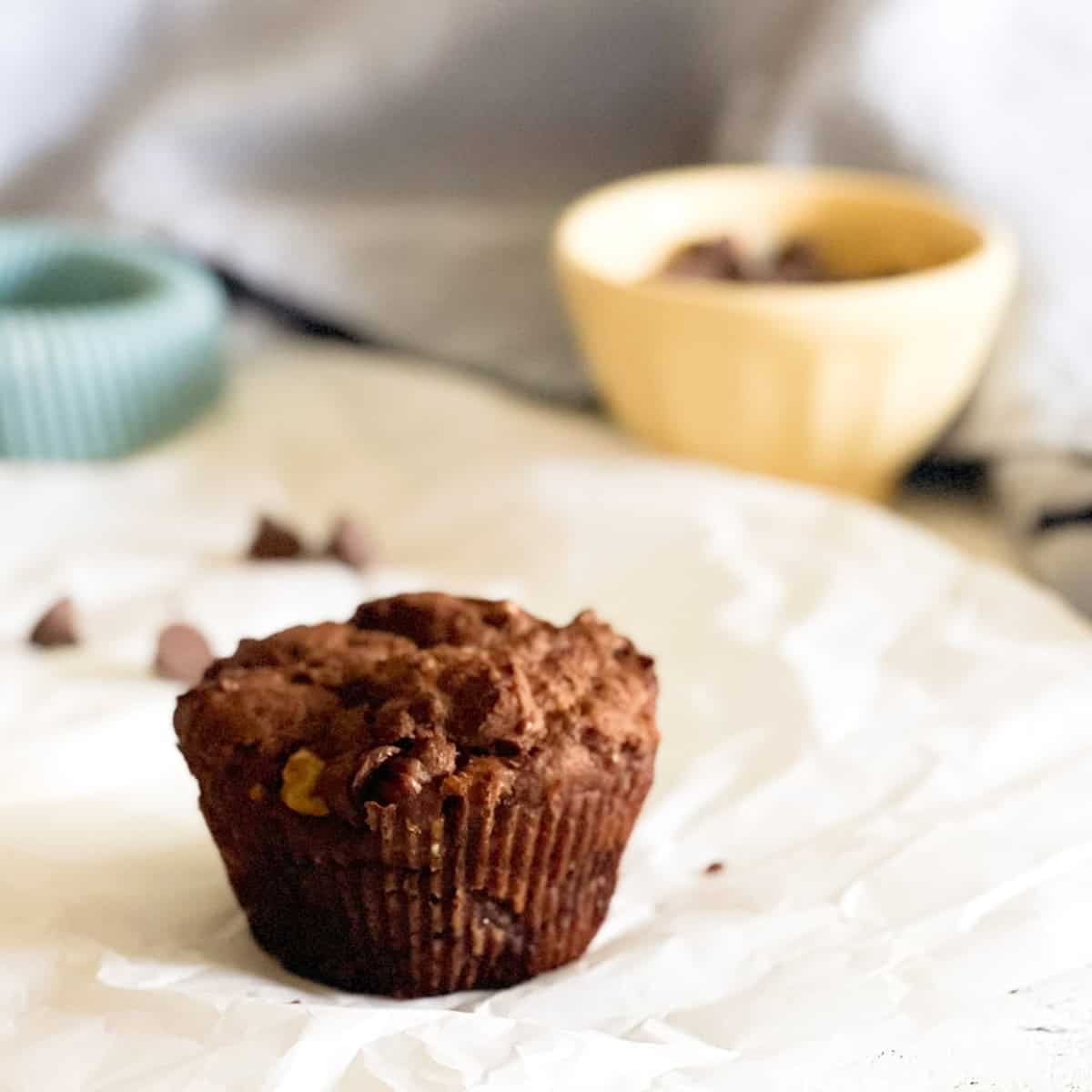 A chocolate chip muffin sits on a crumpled sheet of parchment paper, with a yellow bowl and green ramekin blurred in the background.